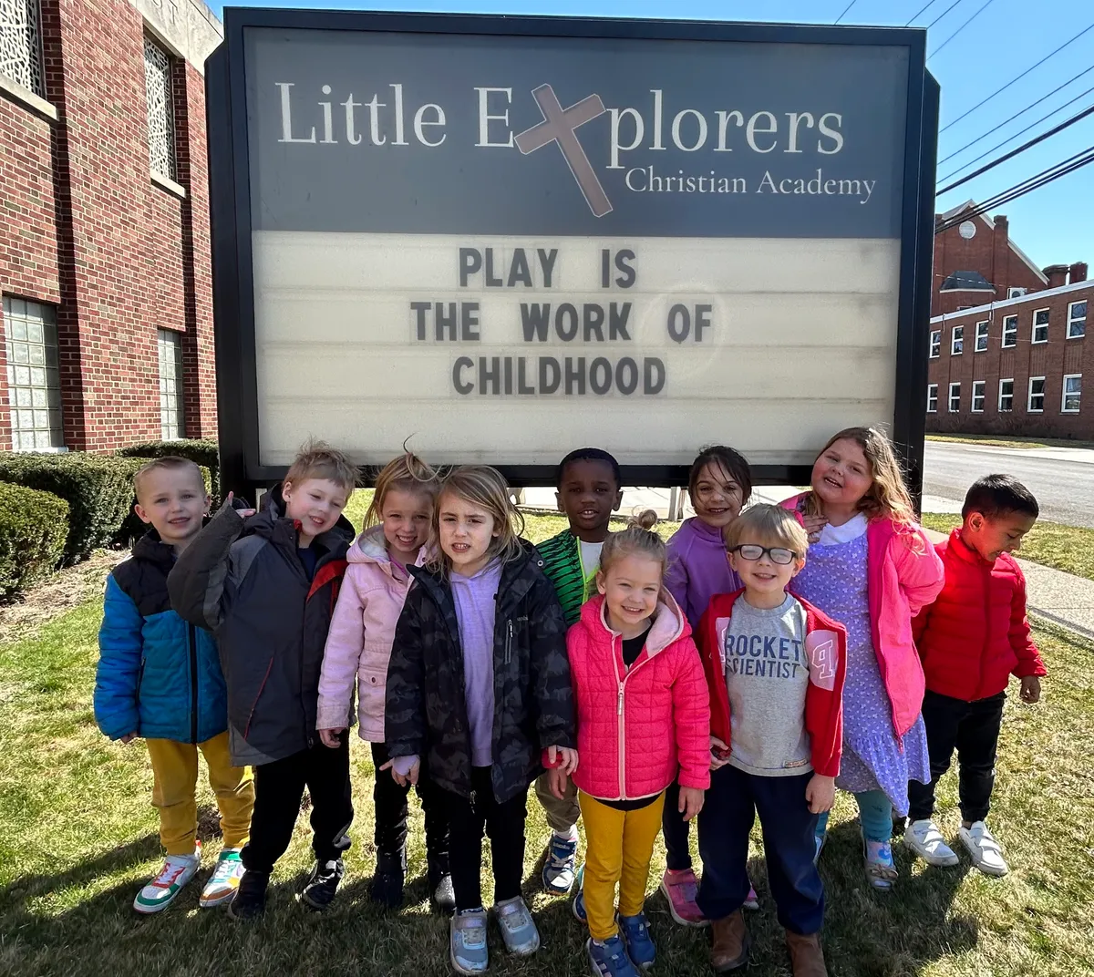 Little Explorers students gathered in front of the school sign reading 'Play is the work of childhood'